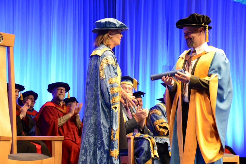 Max Murdo standing on the stage in his ceremonial graduation robes, receiving a scroll from a lady also in ceremonial robes